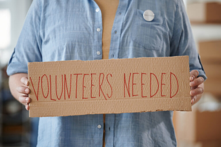 Person holding a sign reading Volunteers Needed to encourage community support.